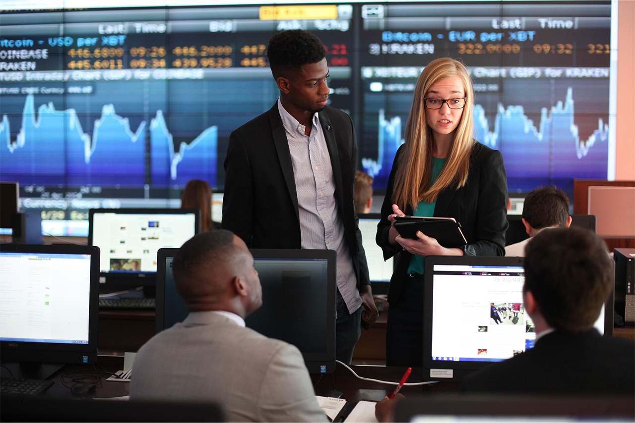 Four business students discuss data on a tablet in trading room displaying current stocks on the screens within the Becker Business Building.