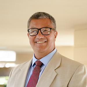 Headshot photo of Tom Curle wearing a beige suit with a red polka dot tie underneath a covered walkway on campus.