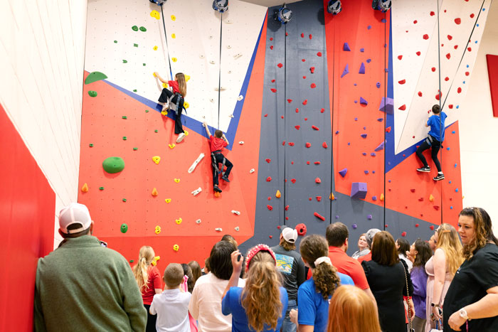 Children climbing rock wall in the gymnasium as parents and others watch below.