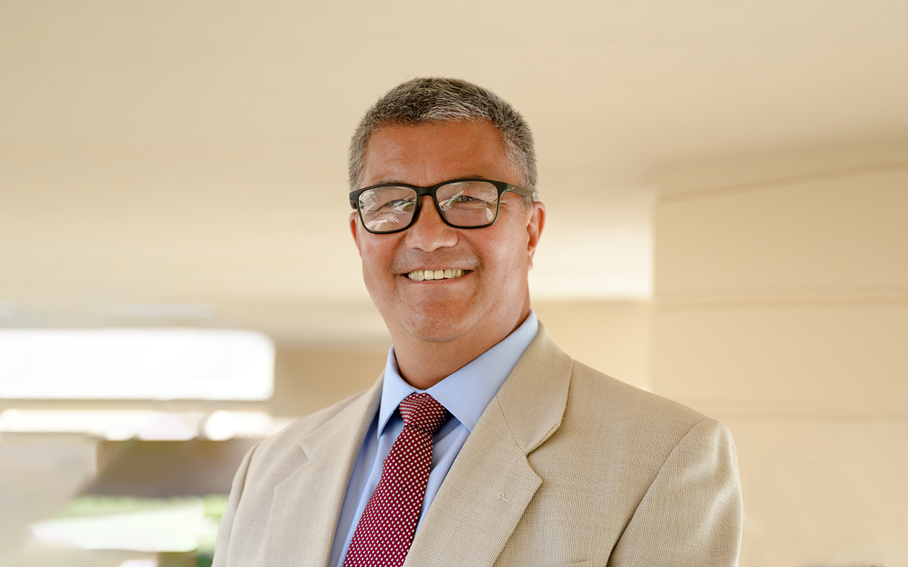 Headshot photo of Tom Curle wearing a beige suit with a red polka dot tie underneath a covered walkway on campus.