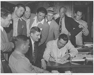 James Haley at a desk with John Ringling. Ringling writes on a piece of paper while Haley and a group of people watch